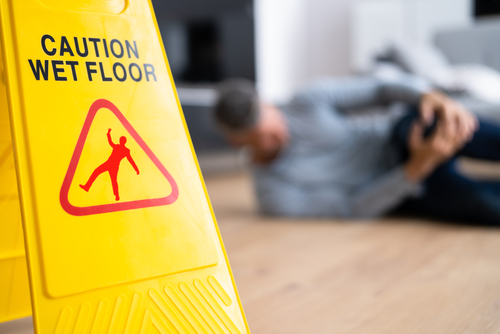 A yellow "Caution Wet Floor" sign is prominently displayed in the foreground, while a person in the background lies on the floor holding their knee, indicating a slip and fall accident.