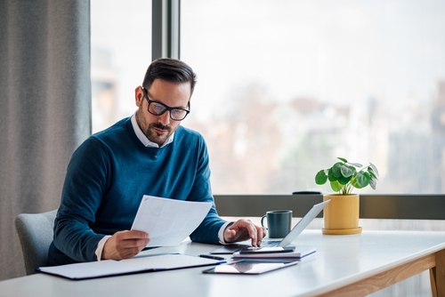 A man in glasses, wearing a blue sweater, is sitting at a desk in a well-lit office. He is reviewing documents while working on a laptop, with a plant and coffee cup on the desk.