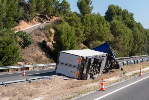 A large truck has overturned on the side of a highway, partially blocking the road, with orange traffic cones placed around the accident site, surrounded by trees and a hillside.