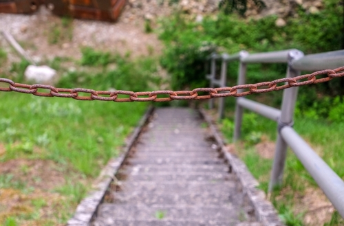 A rusty chain blocks access to a set of outdoor stairs with metal railings, leading down a steep, overgrown path. The setting suggests an unsafe or hazardous condition.