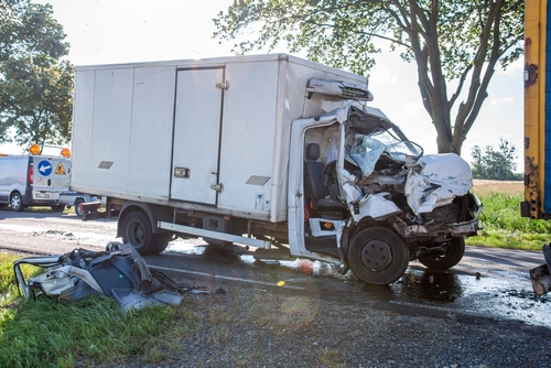 A heavily damaged delivery truck after a serious collision on the road, with debris scattered nearby and emergency responders present in the background.