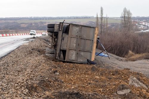 An overturned truck lying beside a rural road under construction, with the truck's cargo spilled and debris scattered around, illustrating a severe vehicular accident in a construction zone.