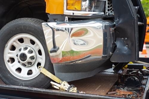 A close-up of a damaged front section of a truck with a dented chrome bumper, secured on a flatbed tow truck, highlighting a vehicle involved in an accident.