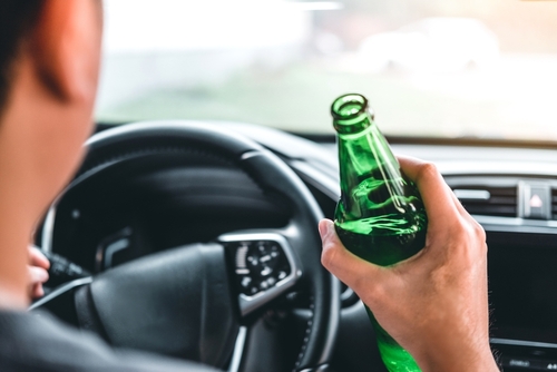 A person holds an open bottle of alcohol while driving, gripping the steering wheel with the other hand, highlighting a dangerous behavior of drinking and driving.