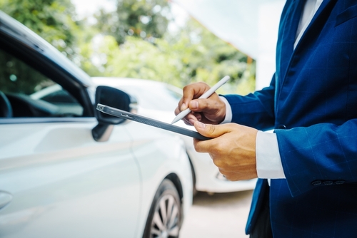 The image depicts a close-up view of a man in a blue business suit, examining or writing on a digital tablet while standing next to a white car. Only his hands and part of his suit are visible, focusing the viewer's attention on the activity with the tablet. The setting appears to be outdoors, with green foliage softly blurred in the background, suggesting a tranquil, professional scenario, likely involving vehicle assessment or documentation.