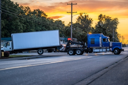 A large blue tow truck is seen towing a white delivery truck during sunset. Power lines, trees, and buildings are visible in the background, and the scene takes place on a road with little traffic.